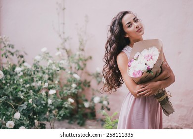Young beautiful woman in a pink dress posing with a bouquet of peonies in a rose garden