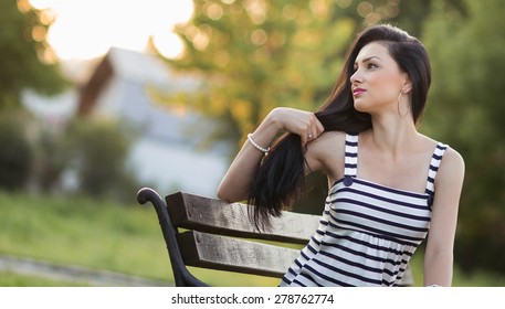 Young Beautiful Woman Playing With Her Long Hair, Outdoors - Outside. Pretty Hispanic Alone Girl Posing On Bench In A Park Looking To Side, Profile - Sideways Concept