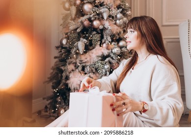 Young Beautiful Woman Plus Size Model In White Knitted Casual Sweater Sits In Living Room Near Christmas Tree And Opens Gift Boxes, Christmas And New Year Concept