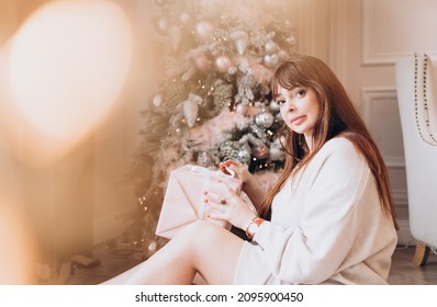 Young Beautiful Woman Plus Size Model In White Knitted Casual Sweater Sits In Living Room Near Christmas Tree And Opens Gift Boxes, Christmas And New Year Concept