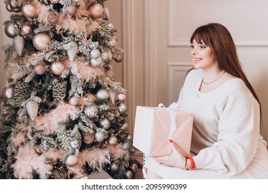 Young Beautiful Woman Plus Size Model In White Knitted Casual Sweater Sits In Living Room Near Christmas Tree And Opens Gift Boxes, Christmas And New Year Concept