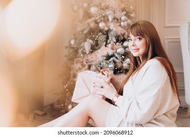 Young Beautiful Woman Plus Size Model In White Knitted Casual Sweater Sits In Living Room Near Christmas Tree And Opens Gift Boxes, Christmas And New Year Concept