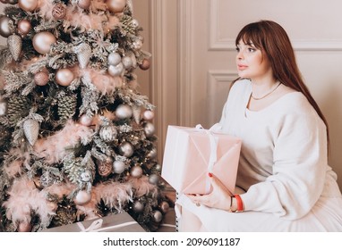 Young Beautiful Woman Plus Size Model In White Knitted Casual Sweater Sits In Living Room Near Christmas Tree And Opens Gift Boxes, Christmas And New Year Concept