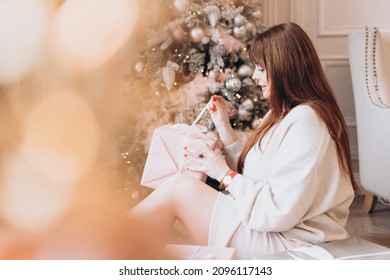 Young Beautiful Woman Plus Size Model In White Knitted Casual Sweater Sits In Living Room Near Christmas Tree And Opens Gift Boxes, Christmas And New Year Concept