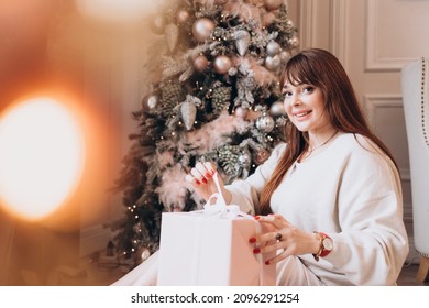 Young Beautiful Woman Plus Size Model In White Knitted Casual Sweater Sits In Living Room Near Christmas Tree And Opens Gift Boxes, Christmas And New Year Concept