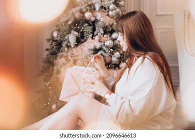 Young Beautiful Woman Plus Size Model In White Knitted Casual Sweater Sits In Living Room Near Christmas Tree And Opens Gift Boxes, Christmas And New Year Concept