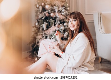 Young Beautiful Woman Plus Size Model In White Knitted Casual Sweater Sits In Living Room Near Christmas Tree And Opens Gift Boxes, Christmas And New Year Concept