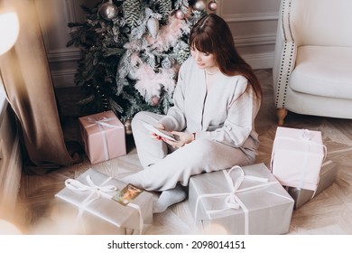 Young Beautiful Woman Plus Size Model In White Knitted Casual Sweater Sits In Living Room Near Christmas Tree And Opens Gift Boxes, Christmas And New Year Concept