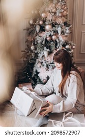 Young Beautiful Woman Plus Size Model In White Knitted Casual Sweater Sits In Living Room Near Christmas Tree And Opens Gift Boxes, Christmas And New Year Concept