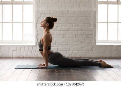 Young Beautiful Woman Practicing Yoga, Lying In Cobra Pose, Doing Bhujangasana Exercise, Attractive Girl In Grey Sportswear, Leggings And Bra Working Out At Home Or In Modern Yoga Studio