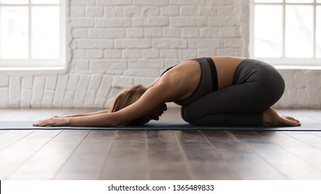 Young Beautiful Woman Practicing Yoga, Lying In Child Pose, Balasana Exercise, Attractive Girl In Grey Sportswear, Leggings And Bra Working Out At Home Or In Modern Yoga Studio With White Walls