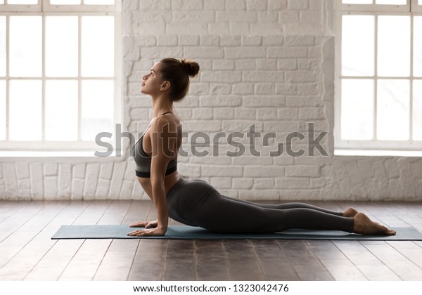 Young beautiful woman practicing yoga, lying in\
Cobra pose, doing Bhujangasana exercise, attractive girl in grey\
sportswear, leggings and bra working out at home or in modern yoga\
studio