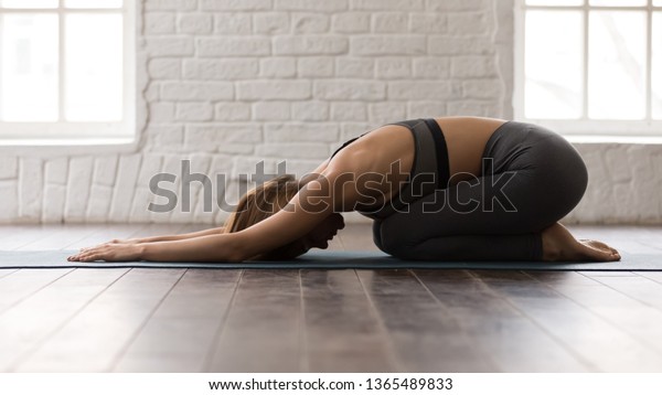 Young beautiful woman practicing yoga, lying in\
Child pose, Balasana exercise, attractive girl in grey sportswear,\
leggings and bra working out at home or in modern yoga studio with\
white walls