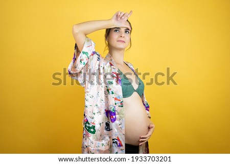 Young beautiful woman pregnant expecting baby wearing pajama over isolated yellow background making fun of people with fingers on forehead doing loser gesture mocking and insulting.