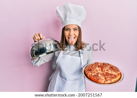 Young beautiful woman professional cook holding italian pizza sticking tongue out happy with funny expression. 
