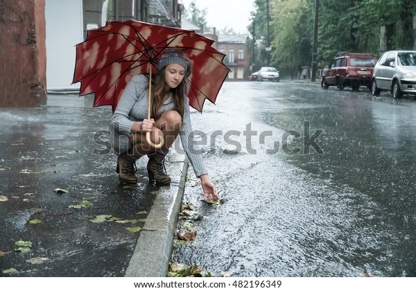 Young beautiful woman in the rain\
under an umbrella leaned over and wets his hand in the\
stream