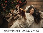 Young beautiful woman reading book, lying near Christmas tree in a cozy decorated room.