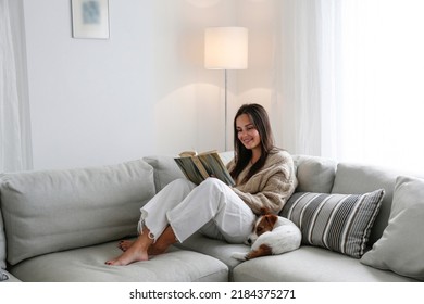 Young beautiful woman reading a book at home with her wire haired jack russell terrier pup. Brunette female and rough coated dog sitting on textile couch. White wall background, copy space, close up.