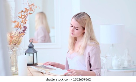 Young Beautiful Woman Reads A Book At The Table In The Living Room. Soft Focus