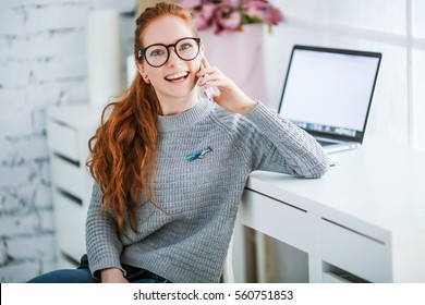 Young Beautiful Woman With Red Hair, Wearing Glasses, Working In The Office, Uses A Laptop And Mobile Phone