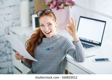 Young Beautiful Woman With Red Hair, Wearing Glasses, Working In The Office, Uses A Laptop And Mobile Phone