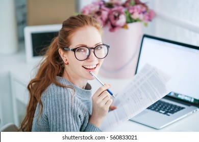 Young beautiful woman with red hair, wearing glasses, working in the office, uses a laptop and mobile phone