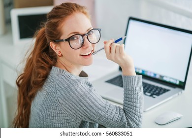 Young Beautiful Woman With Red Hair, Wearing Glasses, Working In The Office, Uses A Laptop And Mobile Phone