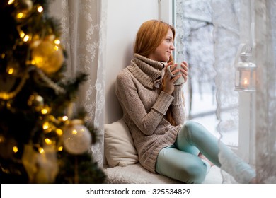 Young Beautiful Woman With Reddish Hair Sitting Home By The Window With Cup Of Hot Coffee Wearing Knitted Warm Sweater. Christmas Tree With Decorations And Lights In The Room, Snowy Winter Outside