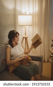 Young Beautiful Woman Relaxing At Home In The Cozy Evening And Reading Book. Fine Art Photo Of A Calm Woman Resting Indoors