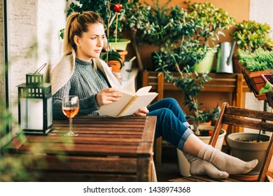 Young Beautiful Woman Relaxing On Cozy Balcony, Reading A Book, Wearing Warm Knitted Pullover, Glass Of Wine On Wooden Table