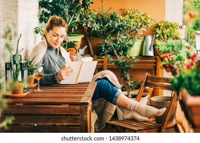 Jovem mulher bonita relaxante na varanda aconchegante, lendo um livro, vestindo pulôver de malha quente, copo de vinho na mesa de madeira