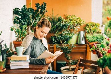 Young Beautiful Woman Relaxing On Cozy Balcony, Reading A Book, Wearing Warm Knitted Pullover, Cup Of Tea Or Coffee On Stack Of Books