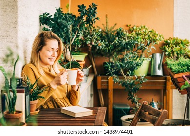 Young beautiful woman relaxing on cozy balcony, reading a book, wearing brown cotton dress, holding cup of tea or coffee - Powered by Shutterstock