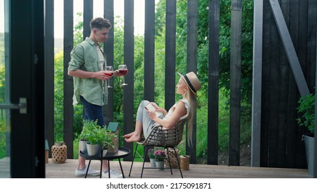 Young Beautiful Woman Relaxing On Cozy Terrace, Reading A Book, Her Boyfriend Is Bringing Her Glass Of Wine, Digital Detox And Weekend Away Concept.