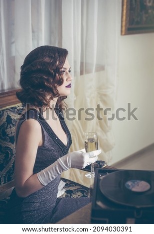 Young beautiful woman in retro style near a vinyl record player with champagne. Vintage interior. Studio photo.