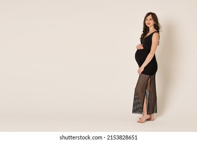 A Young Beautiful Woman In The Second Trimester Of Pregnancy On A Gray Background. A Pregnant Woman In A Full-length Black Dress With Hands On A Round Stomach. The Concept Of Waiting For A Child.