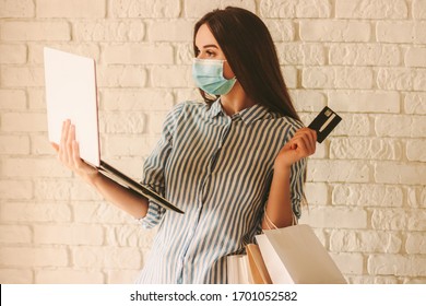 Young beautiful woman shopper in protective face mask hold laptop, credit card and shopping bags in hand. Happy girl in medical face mask shopping at online store. Coronavirus COVID-19, sale, discount - Powered by Shutterstock