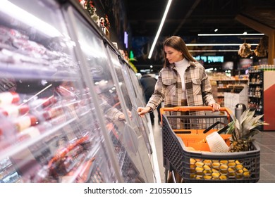 Young Beautiful Woman With A Shopping Cart In The Sausage Department Of A Supermarket Opens The Refrigerator With Products