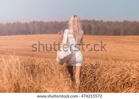 Young beautiful woman in a short white dress with a bouquet of wild flowers walking on the field. At sunset. Outdoors.