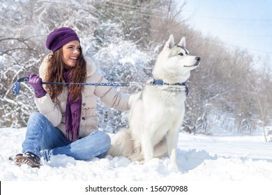 Young Beautiful Woman Sits With Husky Dog In The Winter Forest