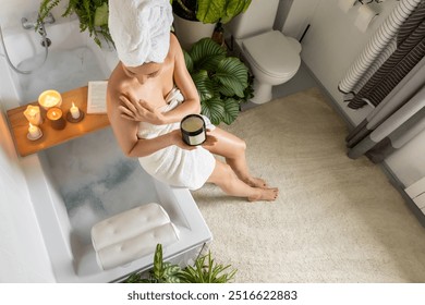 A young, beautiful woman sits on the edge of a bubble bath and applies moisturizer to her skin. Candles, book and house plants. Relax and spa concept at home. Top view. - Powered by Shutterstock