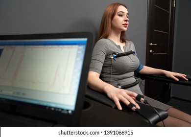 Young Beautiful Woman Sitting With Attached Sensors On Fingers, Hands And Body, Testing With Computer Polygraph, Looking Away. Graph And Polygraph  On Screen Of Lie Detector.