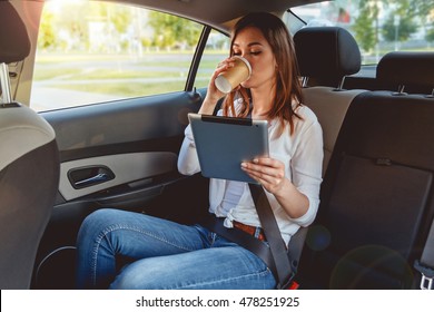Young, beautiful woman sitting in the back seat of the car with a tablet in hand and drinking coffee