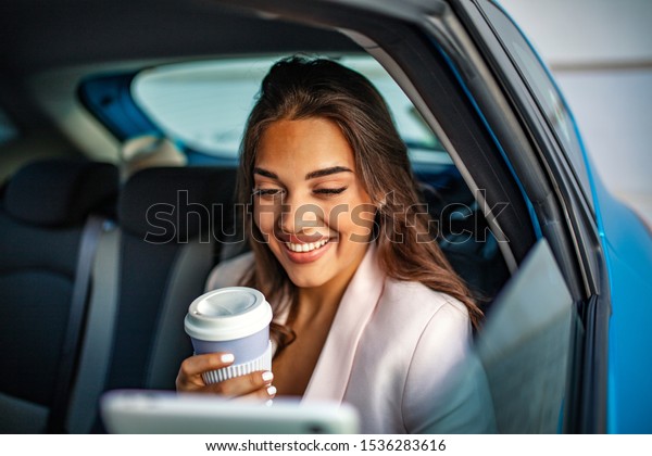 Young, beautiful woman\
sitting in the back seat of the car with a tablet in hand and\
drinking coffee. Female enterpreneur with coffee watching podcast\
on tablet PC