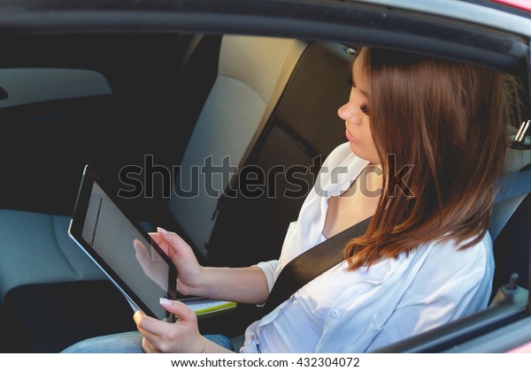 Young, beautiful woman sitting in the back seat of\
a car with a tablet in\
hands