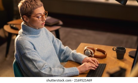 Young Beautiful Woman is Sitting Behind the Table Using Desktop Computer in Sunny Stylish Loft Apartment. Creative Designer Wearing Cozy Blue Sweater and Glasses. Urban City View from Big Window.