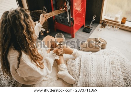 Young beautiful woman sitting by the fireplace in white sweater, drinking wine in cozy log cabin.