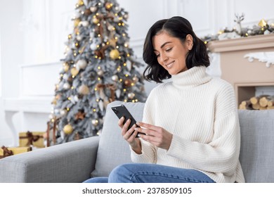 Young beautiful woman sitting near decorated Christmas tree in living room on sofa, smiling and using phone, typing message on smartphone, celebrating new year and Christmas.