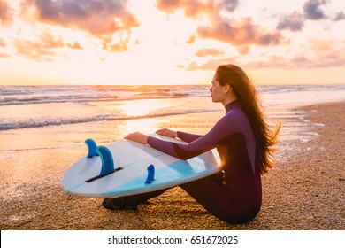 Young Beautiful Woman Is Sitting On The Beach. Surf Girl With Surfboard On Beach At Sunset Or Sunrise. Surfer And Ocean