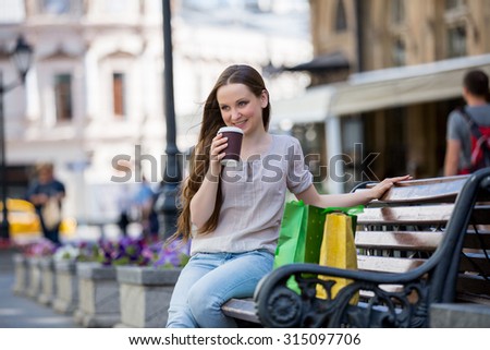 Young beautiful woman sitting on a bench on the street with coffee cup in hand and shopping bags. Drinking coffee and smiling. 
